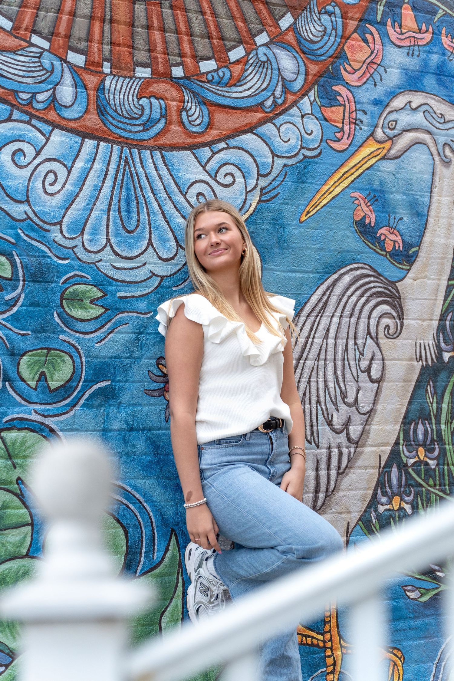 Young blonde woman in white ruffled top and jeans poses smiling in front of a colorful heron mural.