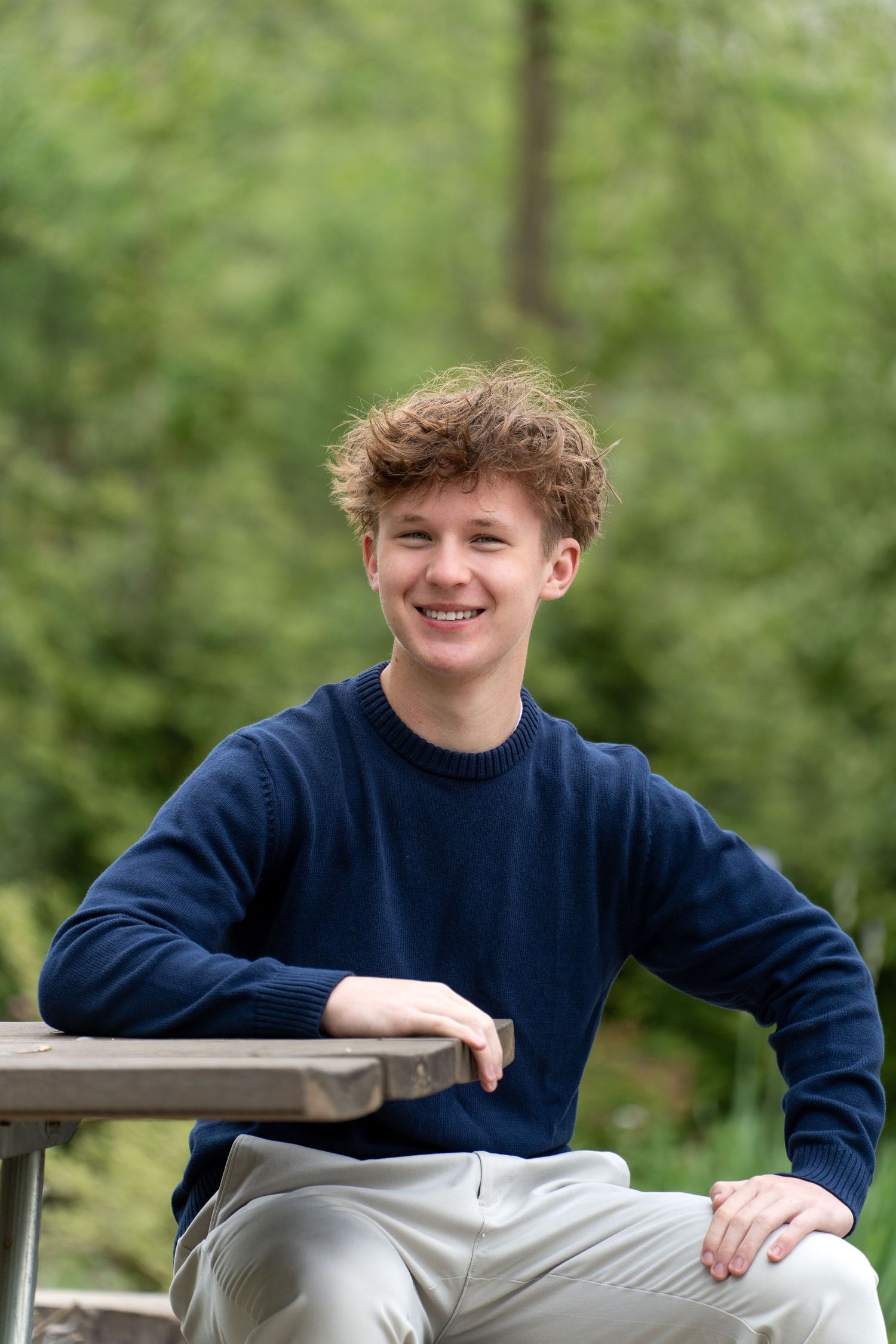 A young person in a navy blue sweater sits casually on an outdoor bench with lush green foliage in the background.