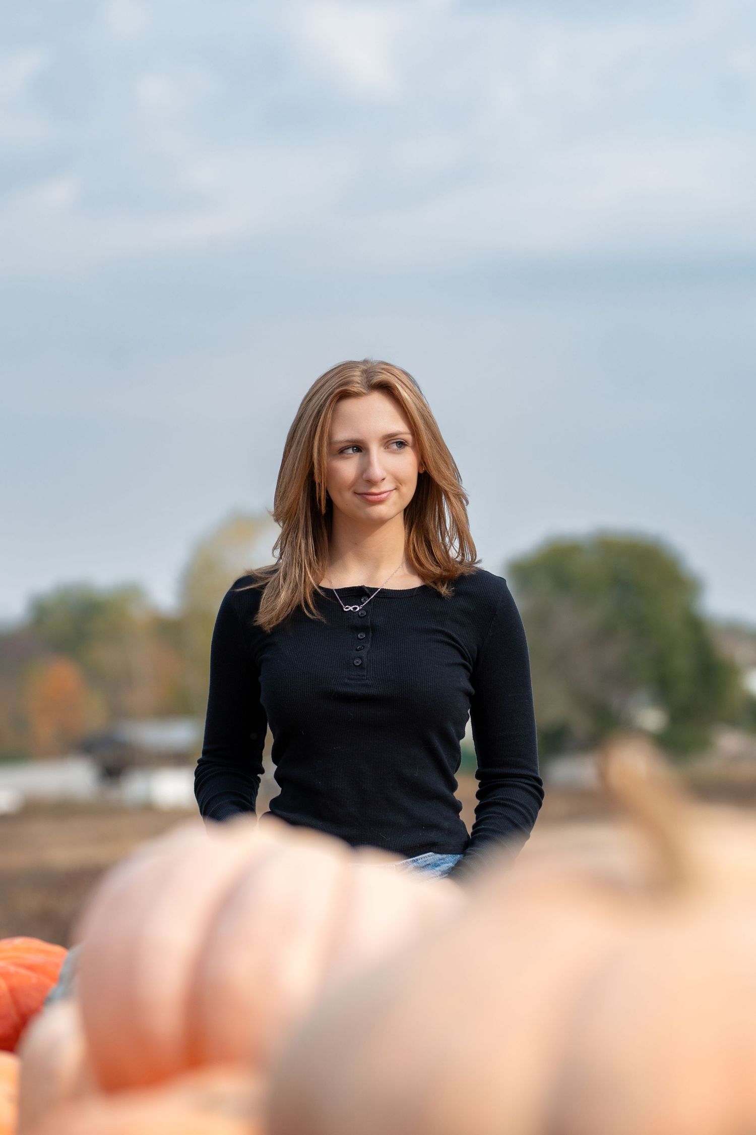 A person in a black long-sleeve top stands outdoors with an autumn pumpkin patch scene in the foreground.
