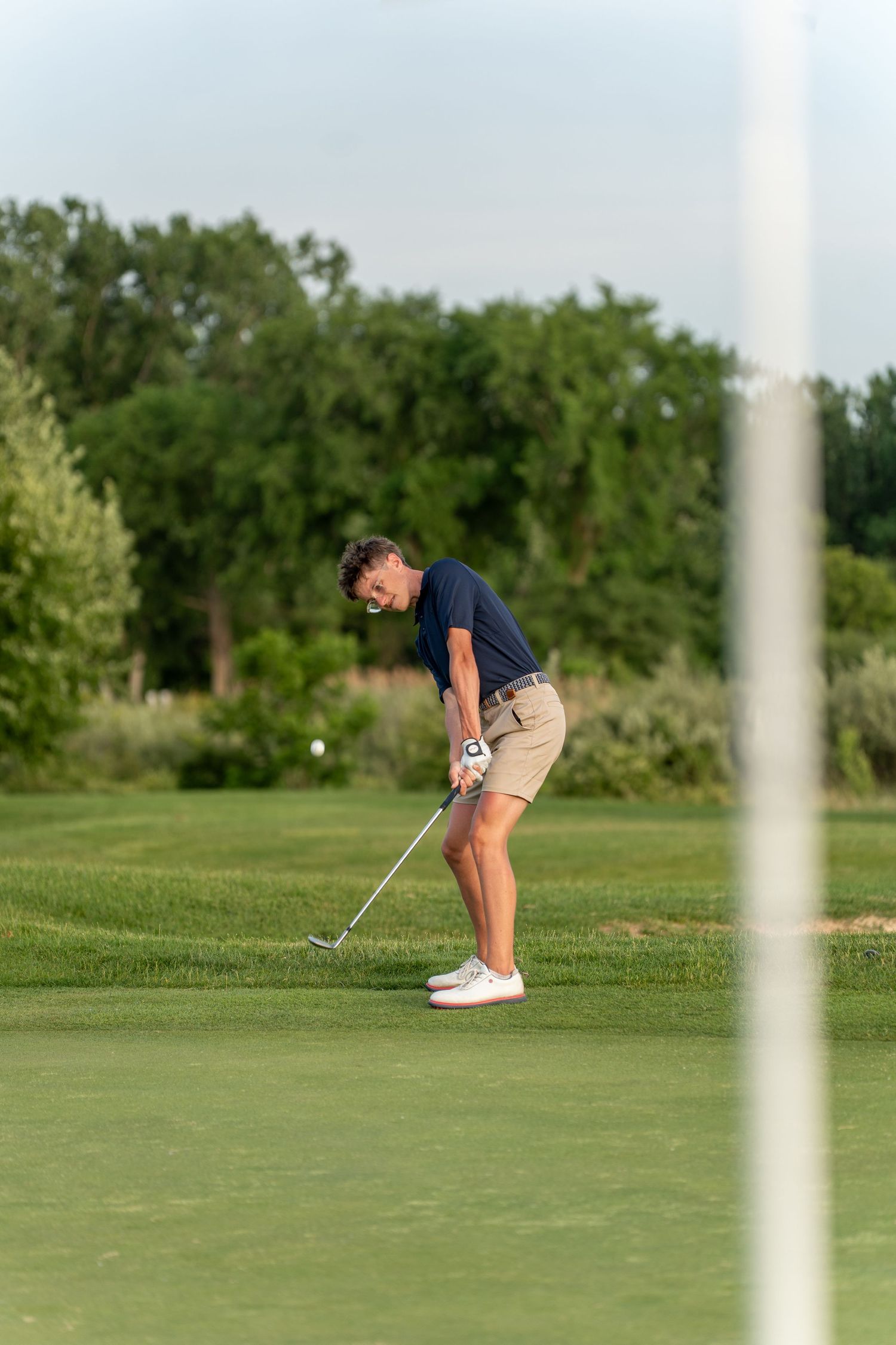 A male golfer in khaki shorts chips a golf ball on a lush green course with trees in the background.
