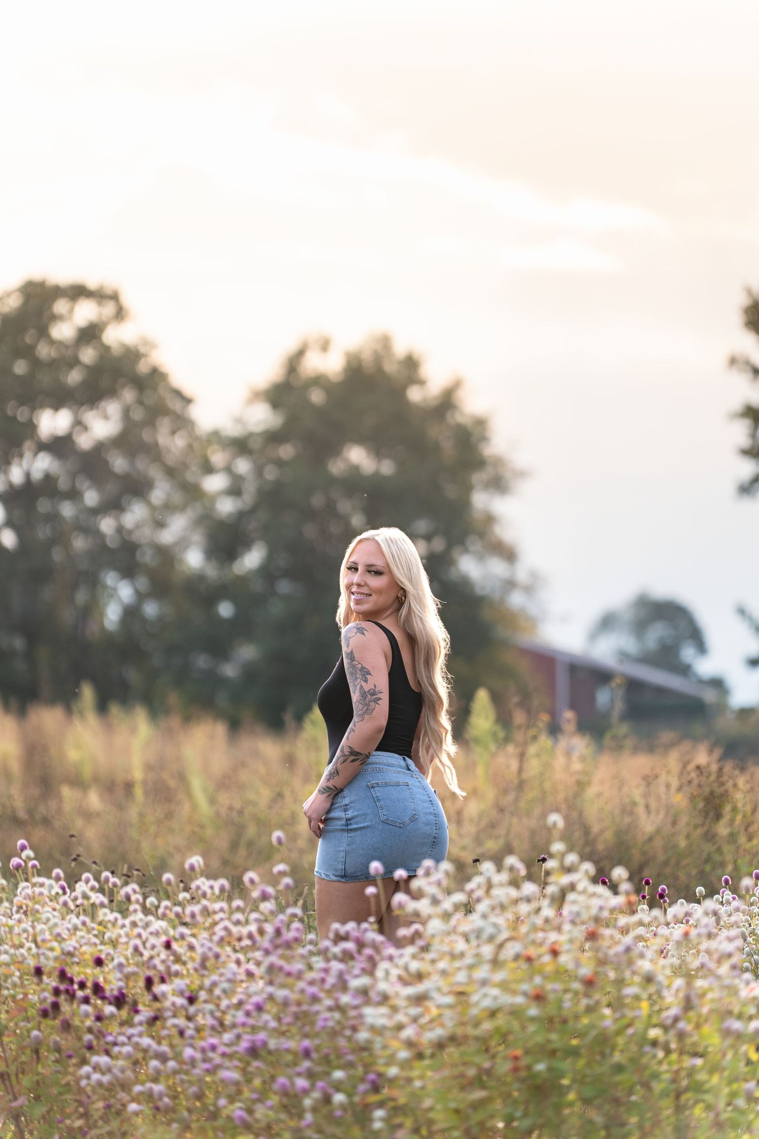 A person in denim shorts stands in a wildflower meadow at sunset with tall grass and flowers surrounding them.