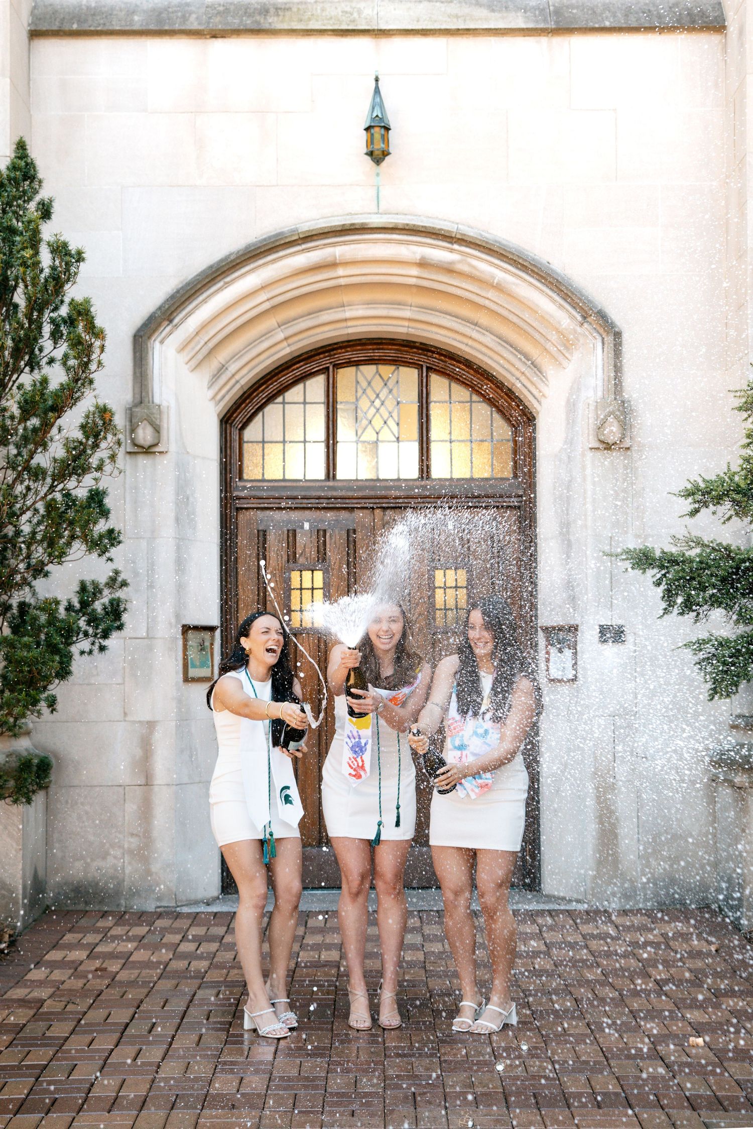 Three women in white dresses celebrate by popping champagne bottles in front of an elegant arched doorway.