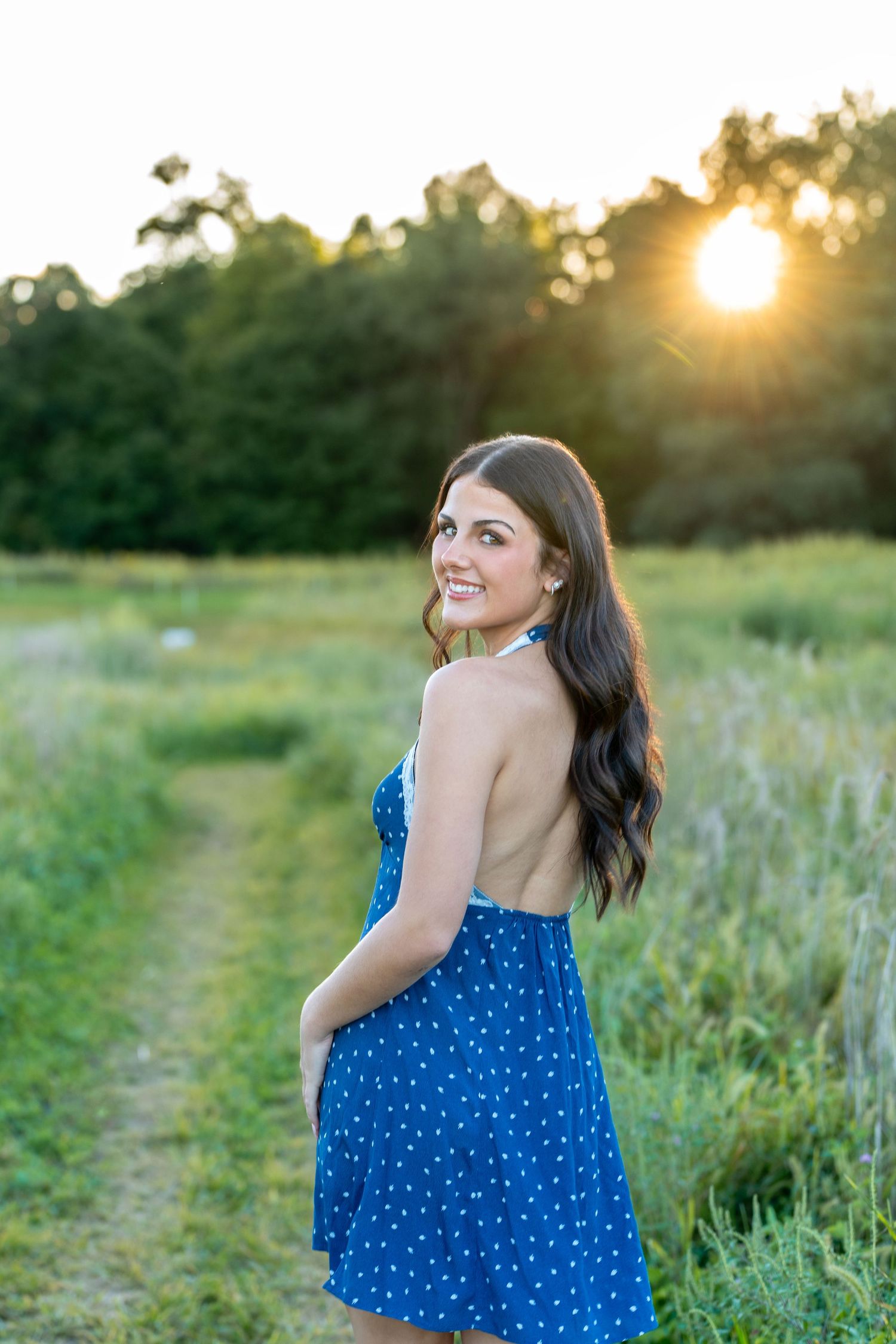 A woman in a blue polka dot dress poses in a sunlit meadow at golden hour.
