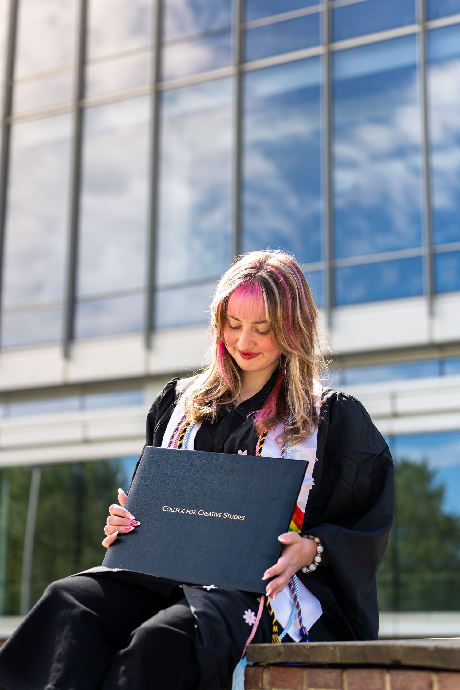 Young woman in graduation gown holding College for Creative Studies diploma, sitting outdoors by modern glass building.