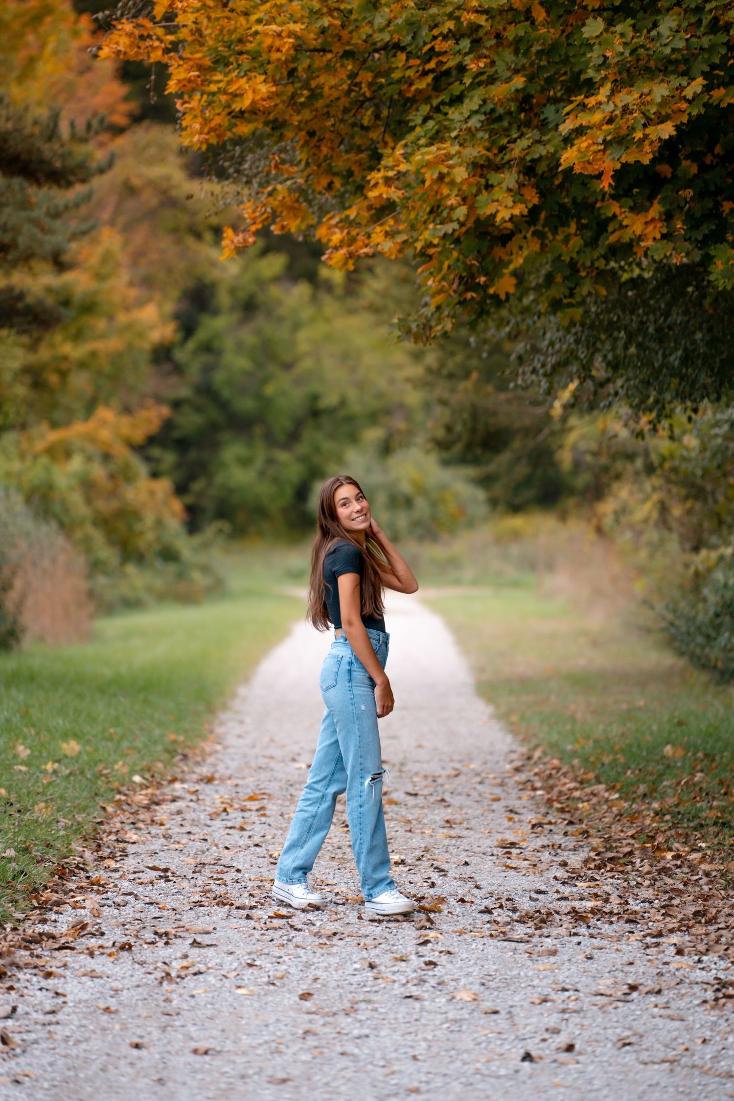 Someone walks along an autumn path beneath golden maple trees wearing jeans and sneakers on a sunny day.