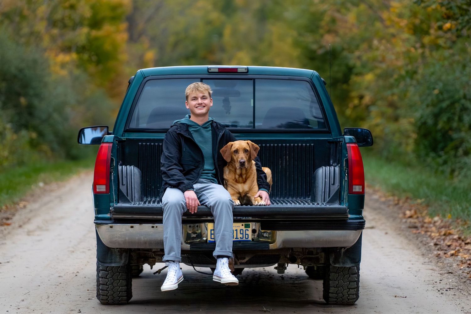 A brown dog and person sit together in the back of a black pickup truck on a fall country road.