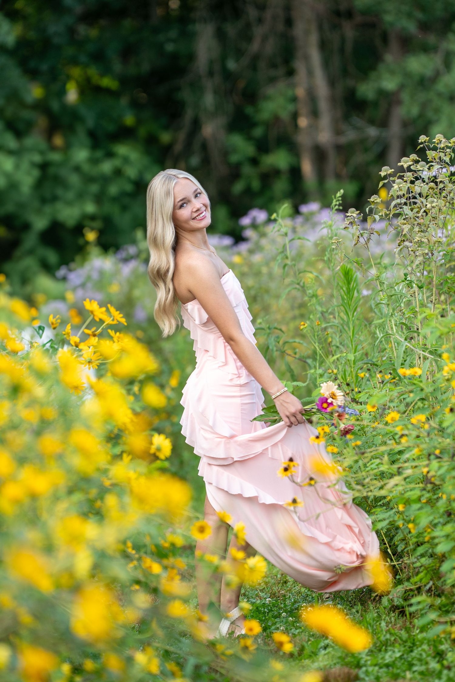 Woman in pink flowy dress dances playfully in a lush wildflower meadow with yellow blooms on a sunny summer day.