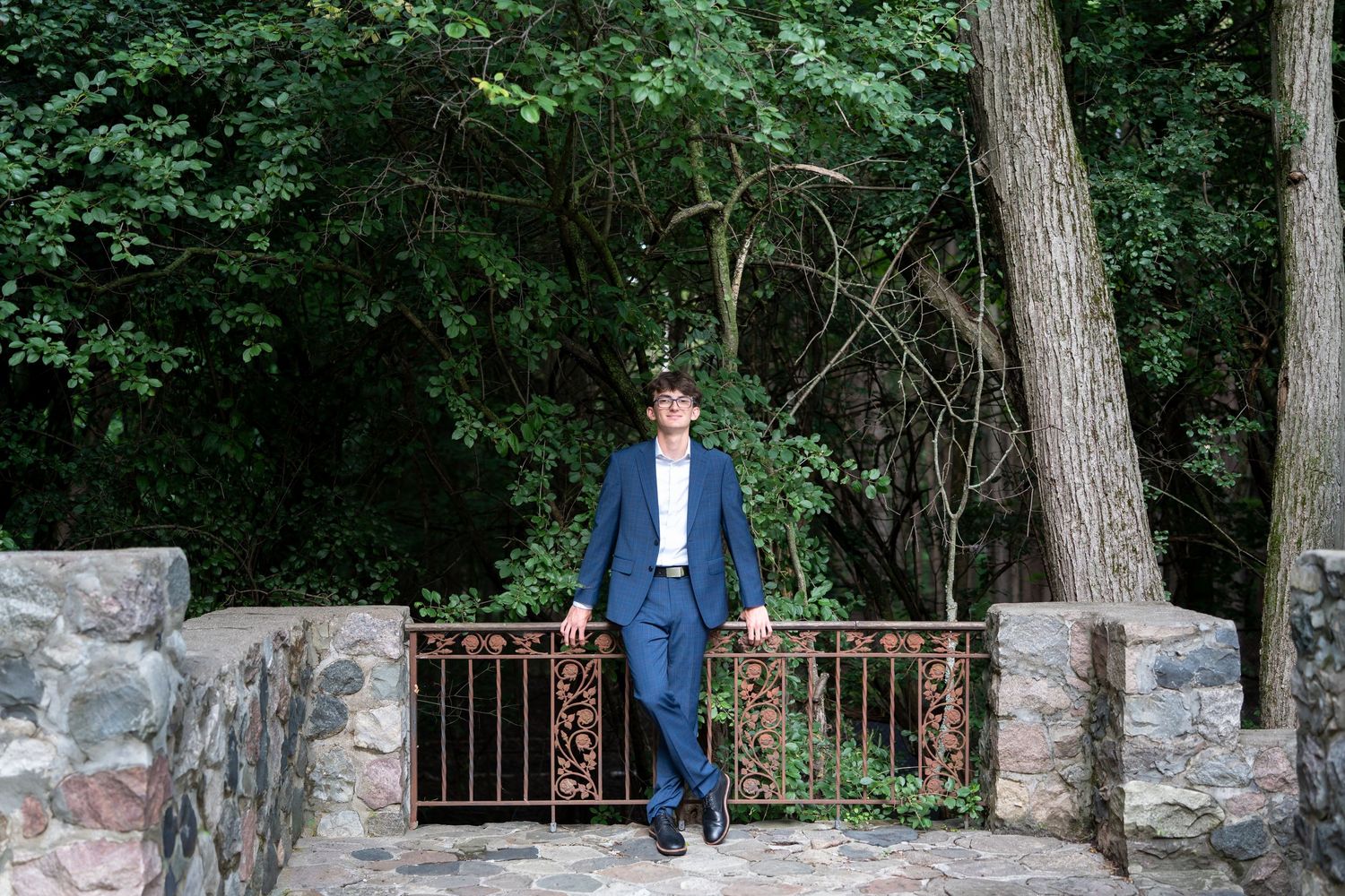 A man in a blue suit leans against an ornate iron fence surrounded by lush green trees in a park setting.