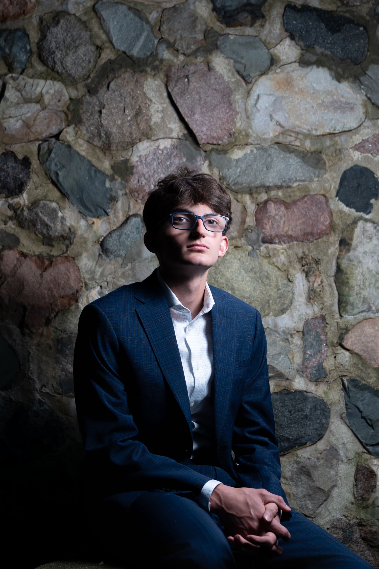 Young man in navy suit and glasses sits against rustic stone wall in dramatic lighting.