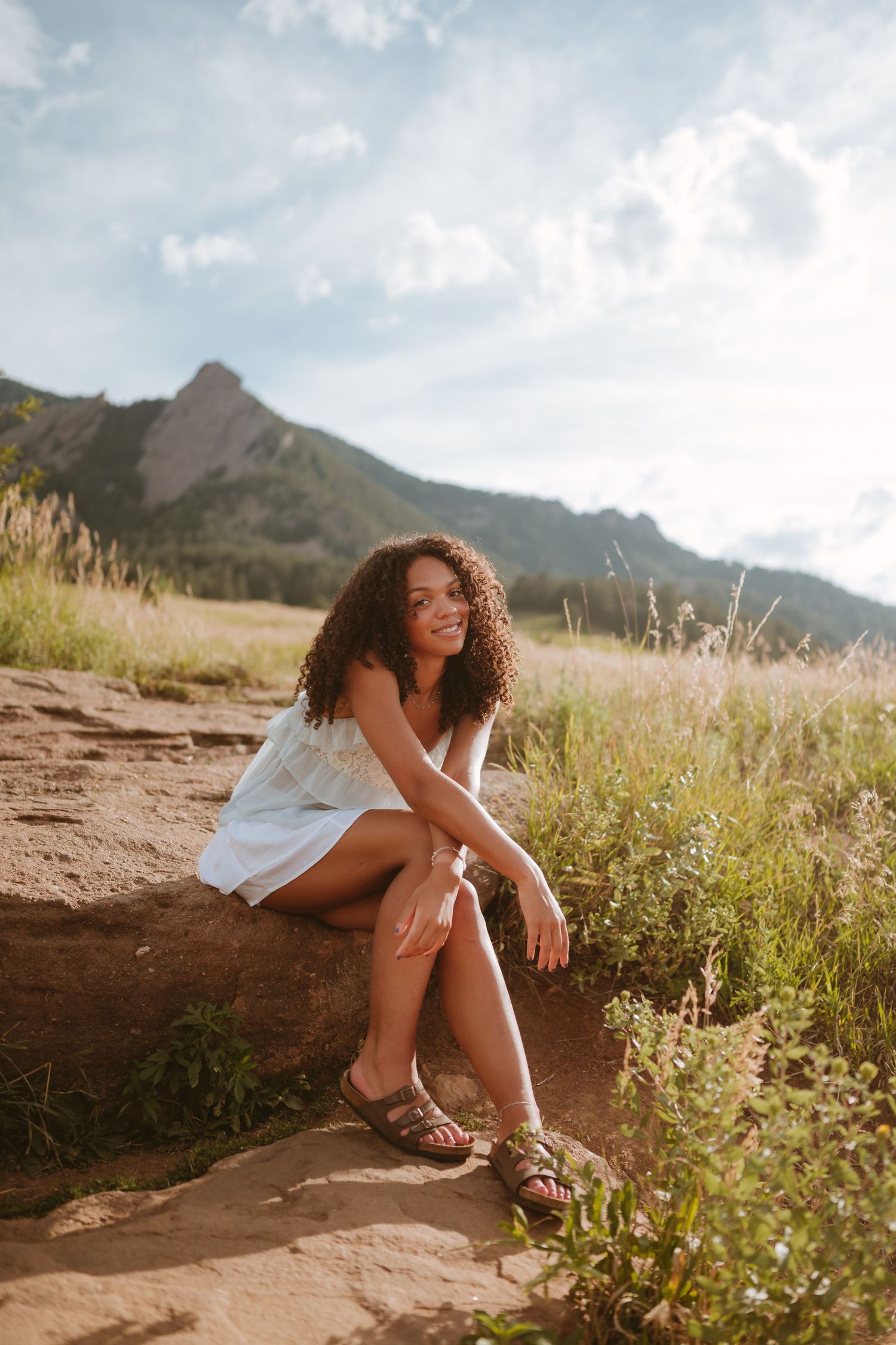 A person in a white dress sits thoughtfully on rocky terrain with mountains and wildflowers in the background at sunset.