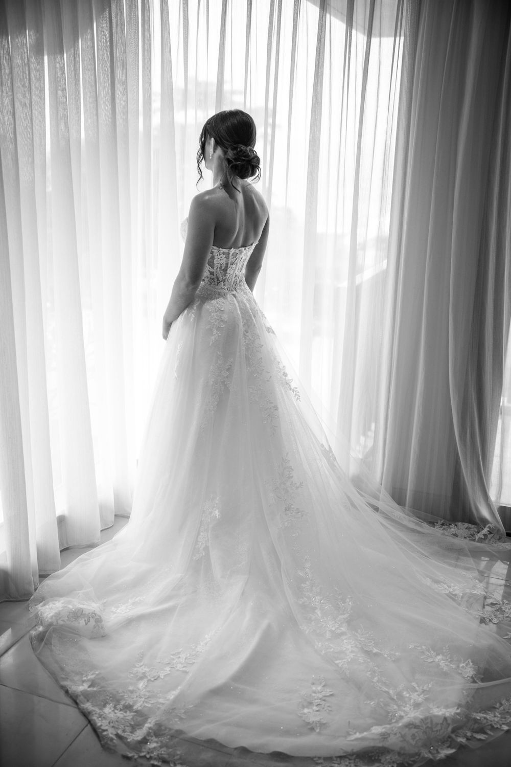 A black and white photograph showing the back of an elegant wedding gown with intricate lace details and a long flowing train.