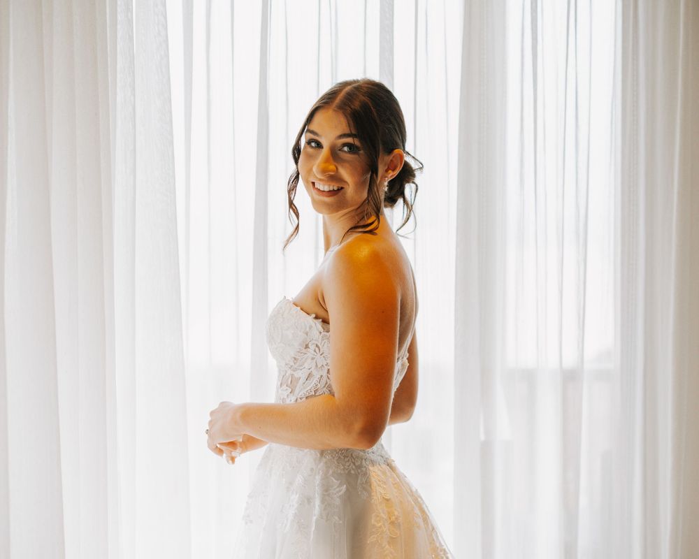 A bride in a strapless lace wedding gown smiles while standing in front of white curtains during golden hour lighting.