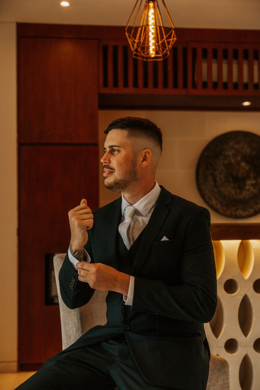 Well-dressed man in dark suit and tie adjusts cuffs in warm-lit room with modern geometric wall decor and lighting.