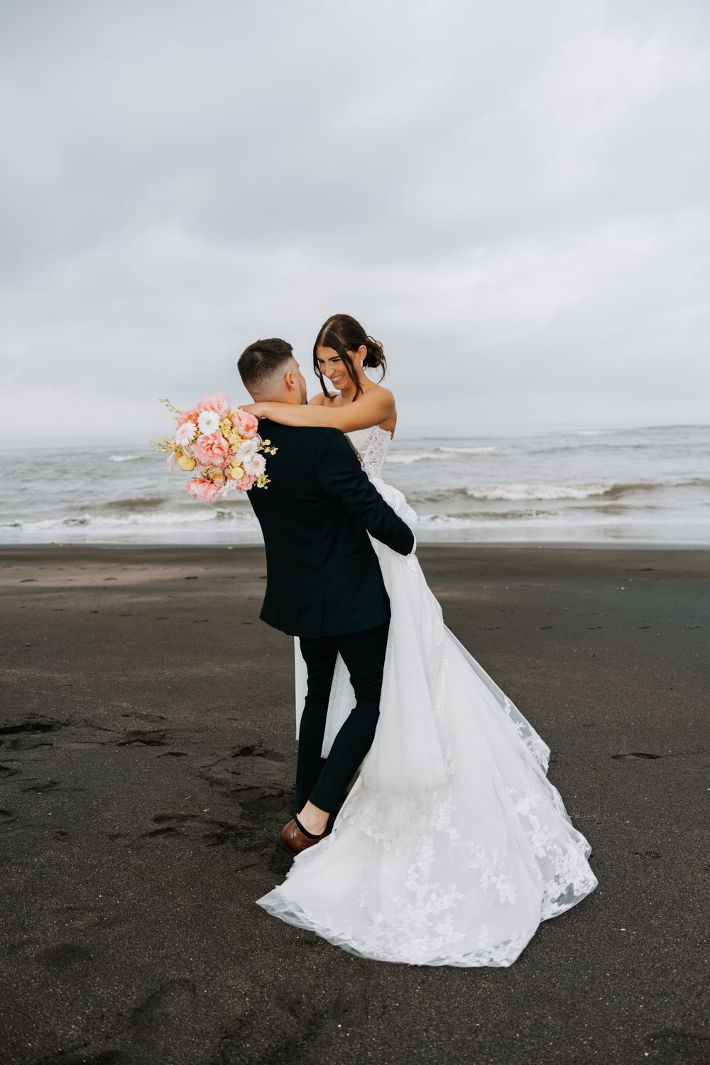 Newlywed couple dancing and embracing on a moody black sand beach with a pink bouquet.