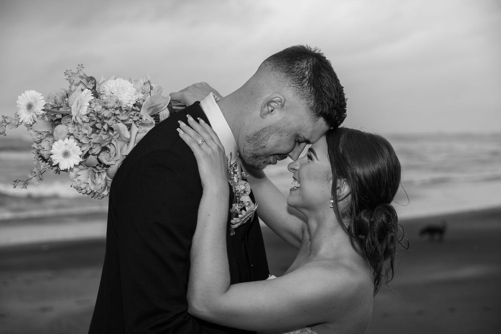 A tender moment between newlyweds embracing on a beach with a bouquet in black and white photography.