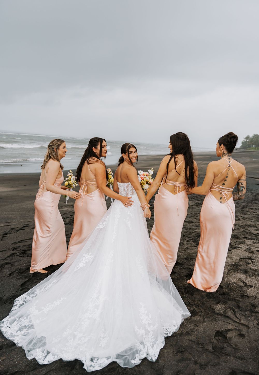 A bridal party wearing blush pink dresses stands together on a moody black sand beach with crashing waves.