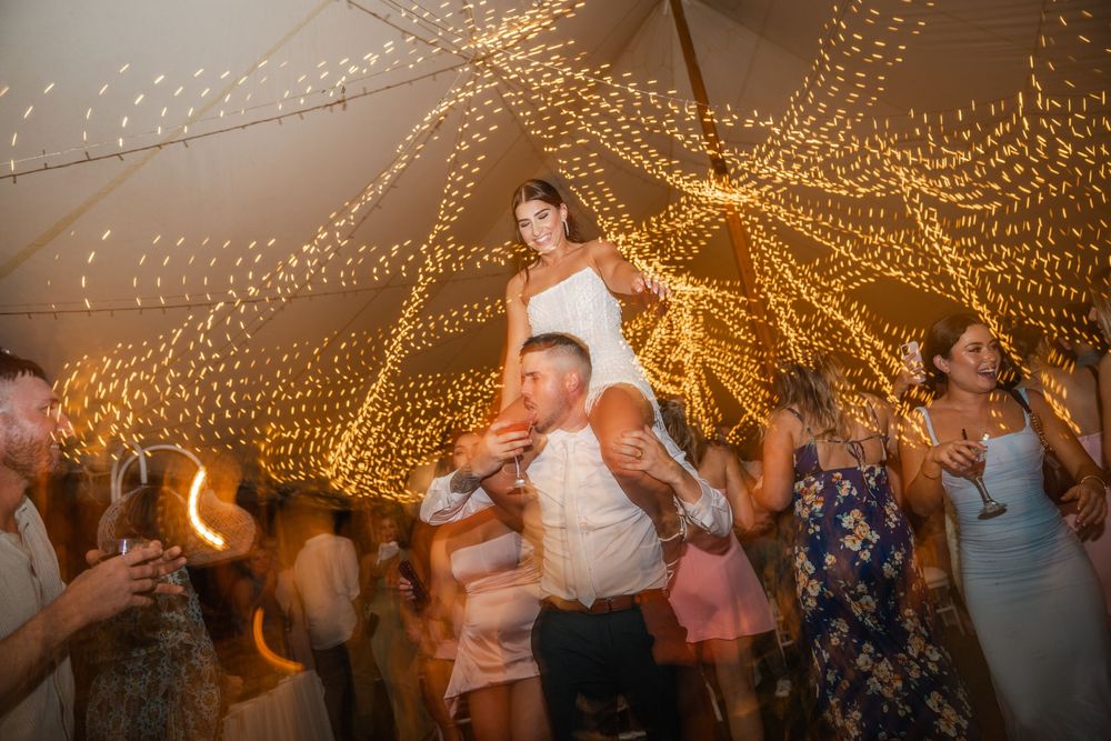 Wedding guests dance together under a canopy of twinkling string lights at an evening reception celebration.