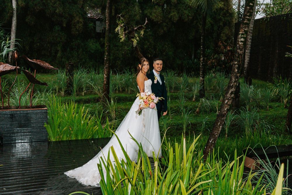 Elegant couple in formal wedding attire poses by tranquil pond surrounded by lush tropical greenery at dusk.