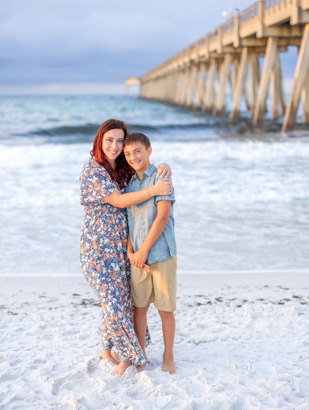 A couple shares an intimate moment at sunset by a wooden pier on a white sandy beach near the ocean waves.