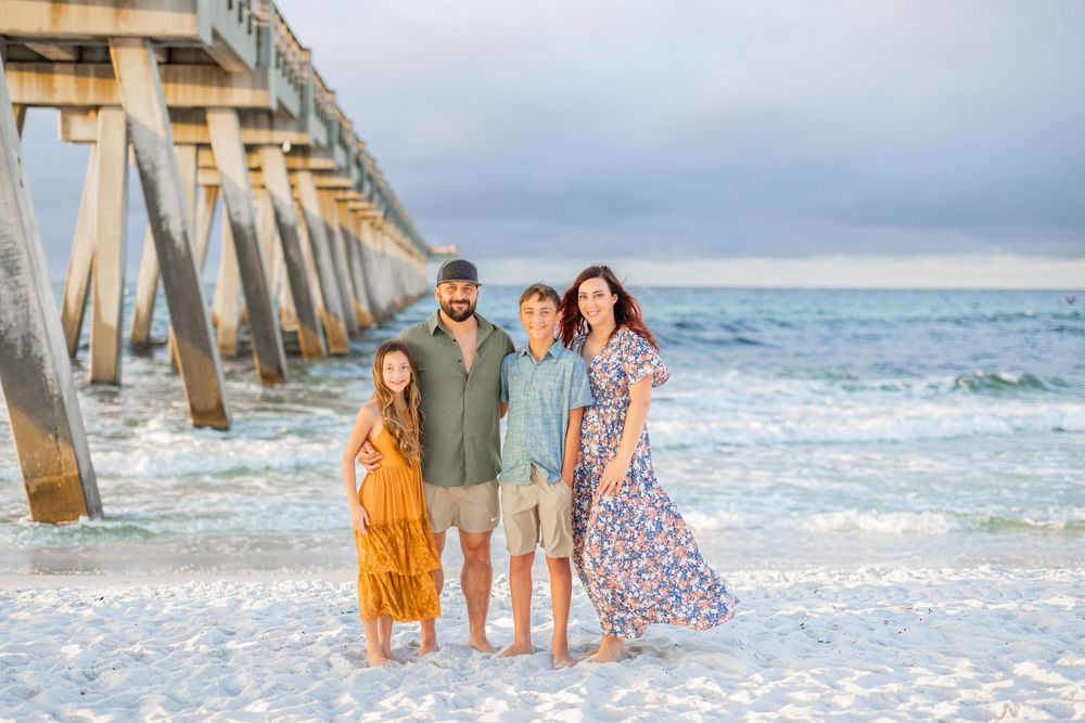 A group stands together on a white sandy beach near a wooden pier with ocean waves in the background.