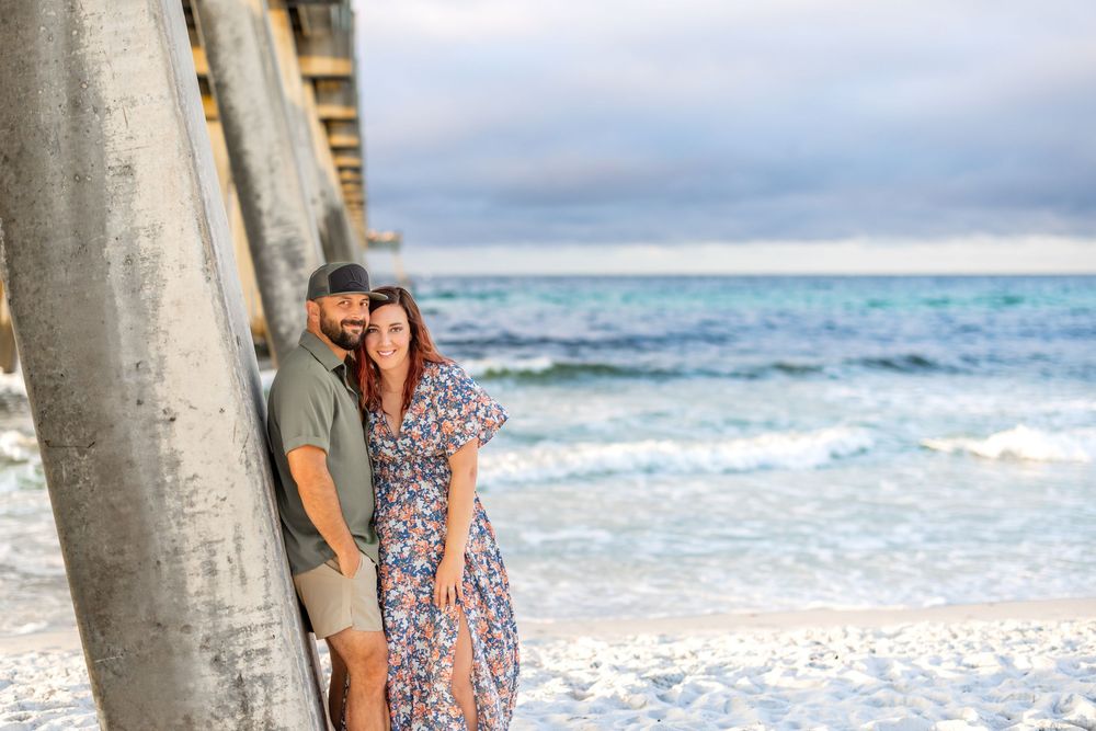 Couple embracing beside wooden pier on sandy beach with ocean waves and cloudy sky in background.