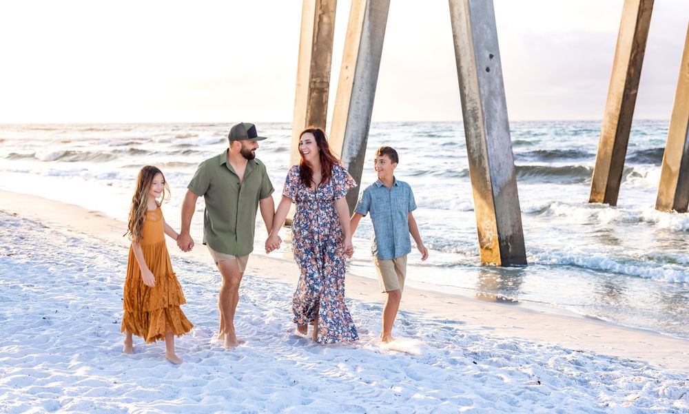 A family walks together barefoot along a white sandy beach near a wooden pier during sunset.