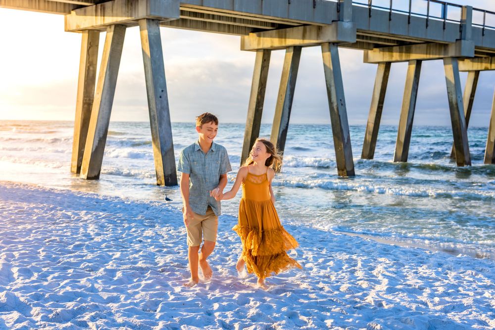 A couple walks along the sandy beach near a wooden pier during sunset while waves roll in.  Sea Glass Photography of Navarre, Fl