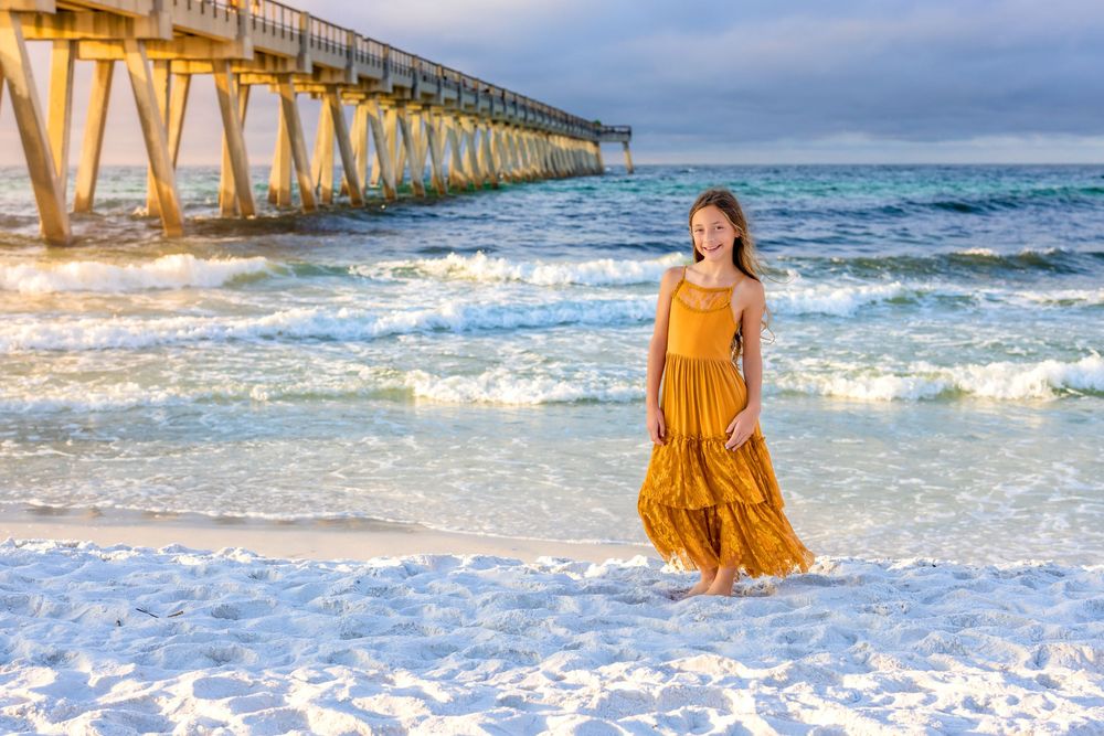 A person in a flowing mustard yellow dress twirls playfully on the beach near a wooden pier at sunset.