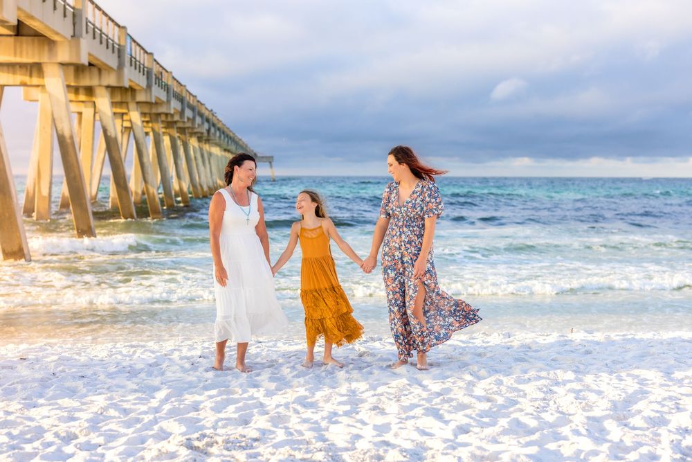 Family walking along a white sandy beach near a wooden pier with waves crashing in the background.