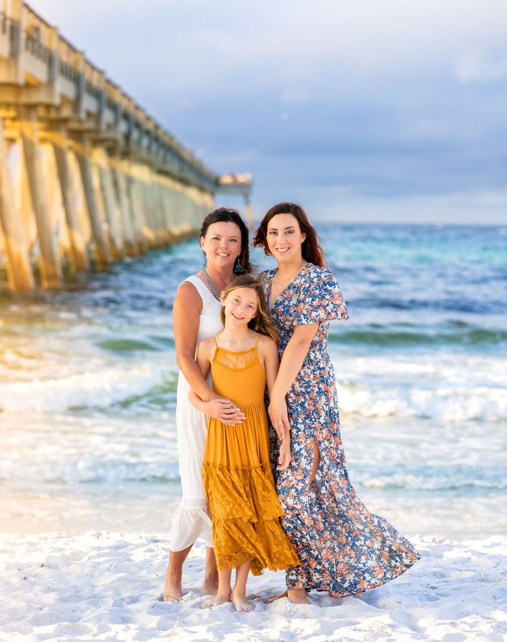 A family poses together on a white sandy beach with a large pier in the background.