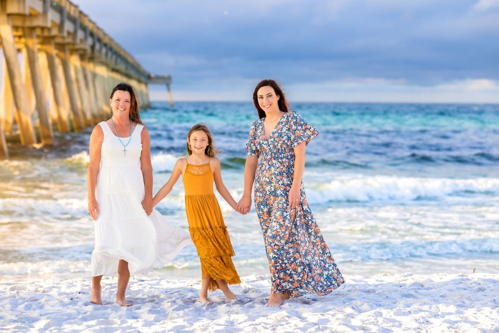 Three people in flowing dresses walk hand in hand along a white sandy beach with a pier and ocean waves in the background.