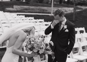 Emotional groom wiping tears as bride holds bouquet during outdoor wedding ceremony with white chairs.
