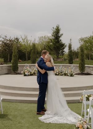 Bride and groom share a tender embrace outdoors at an elegant garden wedding ceremony with floral arrangements.