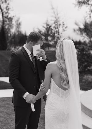 Black and white photo of groom wiping tears while holding brides hand during emotional first look at outdoor wedding.