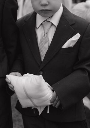 Young ring bearer in black suit holding white pillow at wedding ceremony, black and white photo.