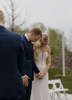 Bride and groom bowing heads in prayer during outdoor wedding ceremony, bride in strapless lace gown, groom in navy suit.