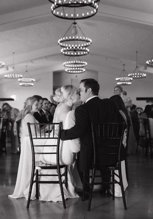 A romantic black and white wedding reception photo showing a couple sharing a kiss while seated in Chiavari chairs.