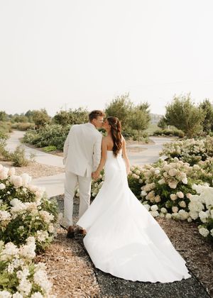 A romantic garden wedding scene with white floral arrangements lining a winding path as a couple shares an intimate moment.