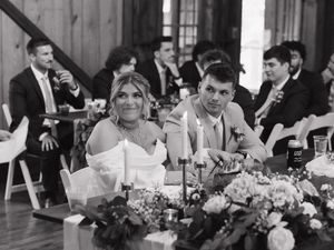 A newly married couple sits at their reception table with floral decorations and candlelight while guests mingle in background.