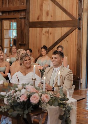 Wedding reception guests seated at long wooden tables in a rustic barn venue share joyful reactions during speeches.