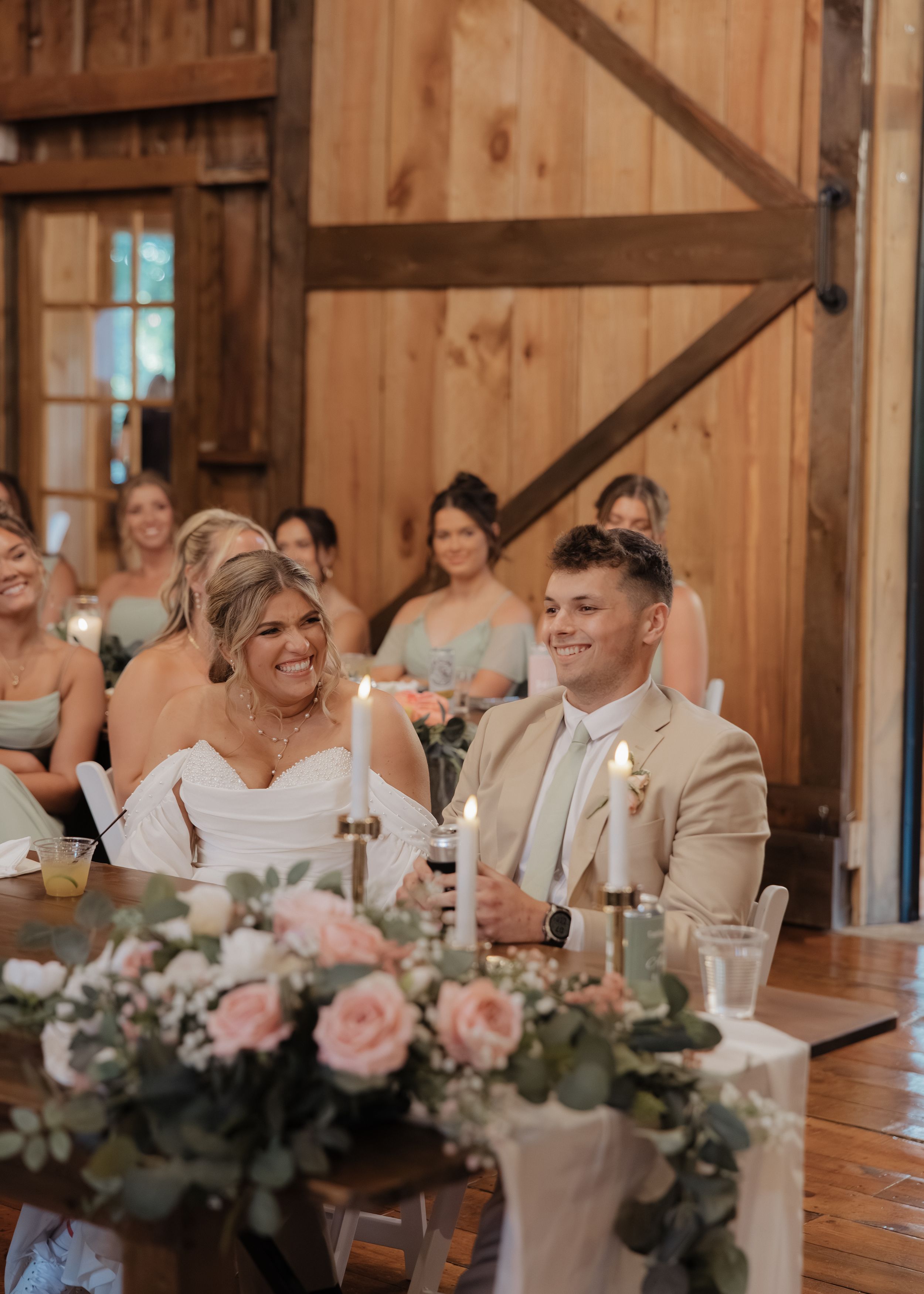 Wedding reception guests seated at long wooden tables in a rustic barn venue share joyful reactions during speeches.