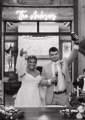 Newlywed couple celebrates with drinks raised high beneath illuminated 'The Arabesque' sign at their wedding reception.