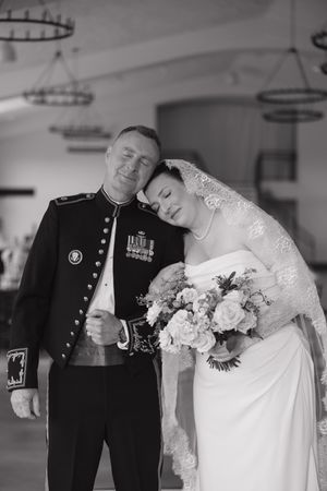 Black and white photo of a bride leaning on a military officer in dress uniform, holding a bouquet at a wedding.