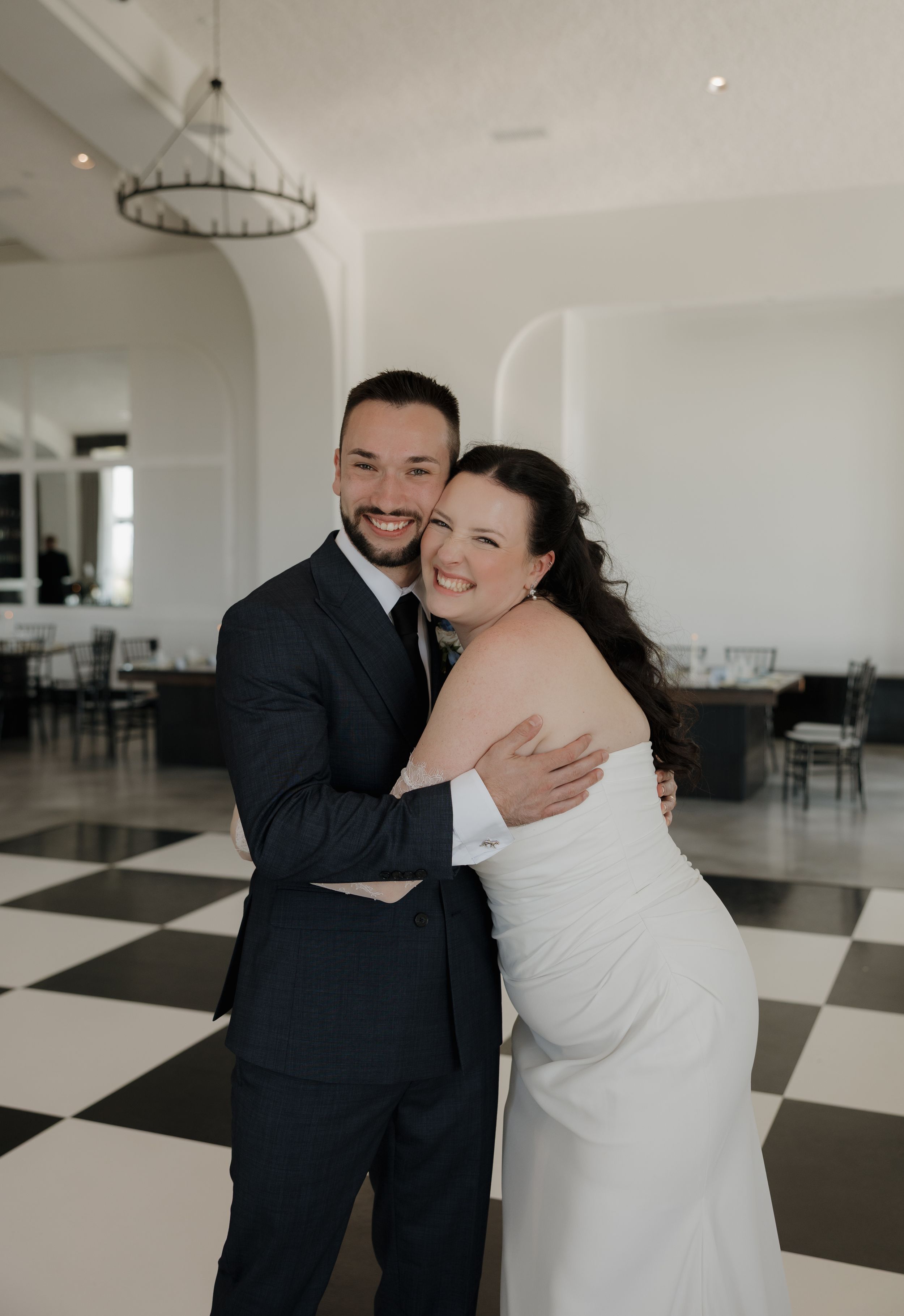 Smiling bride and groom embrace on a black and white checkered dance floor in an elegant white ballroom.