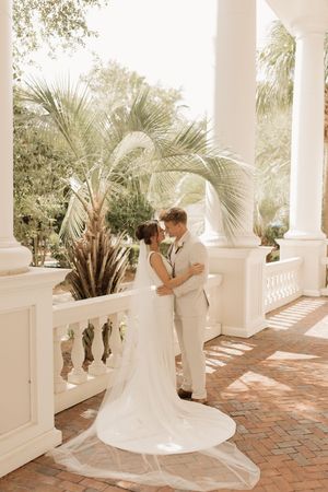 A romantic wedding couple embraces on a sunlit terrace with white columns and tropical palm trees in the background.