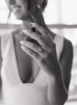Black and white close-up of hands holding a perfume bottle, featuring a wedding ring in soft natural lighting.