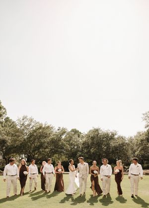 Wedding party members in brown and beige attire stand in a line casting long shadows on grass during golden hour.