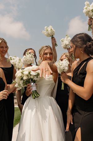 Wedding party in black dresses gathers around bride in white ballgown holding white floral bouquets outdoors.