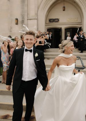 A joyful wedding couple exit their ceremony holding hands while guests celebrate in the church archway background.