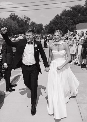 Black and white photo of newlyweds walking down a sidewalk surrounded by guests after their wedding ceremony.